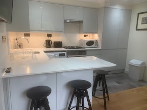 A modern kitchen with white cabinets, a central island with stools, a microwave, and a gas stove. - Kensington Lodge - mins from Natural History Museum, on award-winning Gdn Sq  (London)