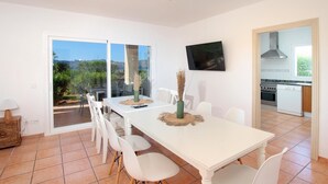 Un'area pranzo con tavolo e sedie bianche, una TV montata a parete e una cucina visibile attraverso una porta aperta.