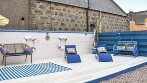 Une terrasse en bois avec des chaises longues, un tapis à rayures bleues et un mur en pierre.