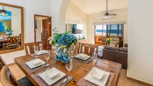 A dining area with a wooden table set for four, a vase with flowers, and a view of the ocean through a window. - Newly Remodeled, Great Views, Beachfront!  (San José del Cabo)