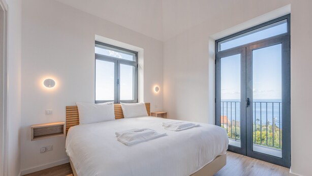 Quarto com cama grande, duas mesas de cabeceira, uma janela e porta para varanda.
