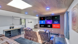 Un salon moderne doté d’un grand téléviseur à écran plat diffusant divers services de streaming, d’un coin salon confortable et d’un design de plafond élégant.