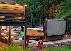 Un área de estar al aire libre con un ambiente rústico, que incluye una mesa y sillas cubiertas, una parrilla portátil y una estructura similar a una carpa decorada con luces colgantes.