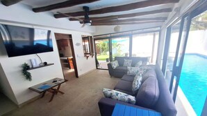 A living room with a sofa, a table, a TV, and a view of the ocean. - Casa Bugambilia-Santa Cruz Pool & walk to Beach! (Avienda)