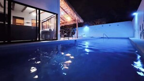 An indoor swimming pool with a glass wall on one side, a thatched roof structure, and a well-lit area with chairs and tables. - Casa Bugambilia-Santa Cruz Pool & walk to Beach! (Avienda)