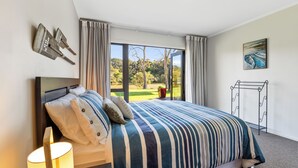 Ein Schlafzimmer mit einem großen Bett, einem Nachttisch, einer Lampe, einem Fenster mit Vorhängen und Blick auf einen Garten.