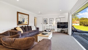 Ein modernes Wohnzimmer mit einer großen Couch, einem Couchtisch und einem Flachbildfernseher. Es gibt einen Balkon mit Blick auf den Garten.