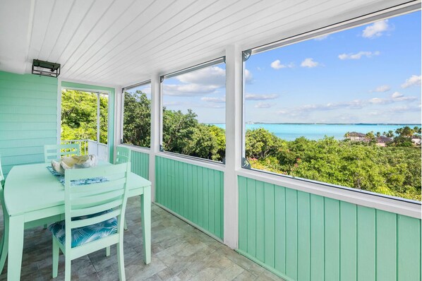 A covered balcony with a table and chairs, large windows offering a view of the ocean and greenery.