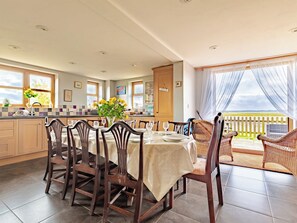 Un coin repas avec une table dressée pour quatre personnes, entourée de chaises en bois, une cuisine en arrière-plan et une vue sur l’extérieur par une fenêtre.