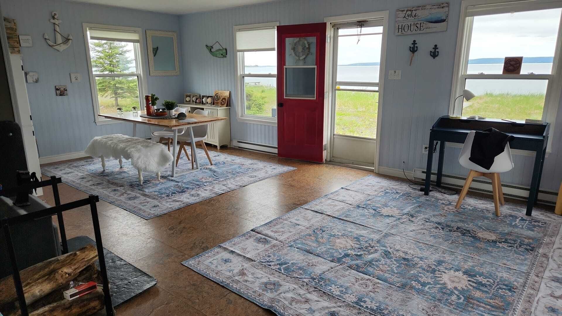 Une pièce avec un plancher en bois, une porte rouge, une table à manger avec des chaises, un piano et une vue sur un lac.