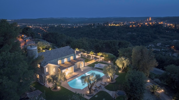 A large, stone house with a swimming pool, surrounded by trees and a view of a distant cityscape at dusk.
