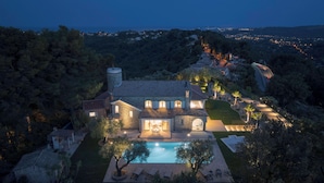 Une grande maison aux murs recouverts de pierres, dotée d’une piscine, entourée d’une végétation luxuriante et offrant une vue sur une ville lointaine au crépuscule.