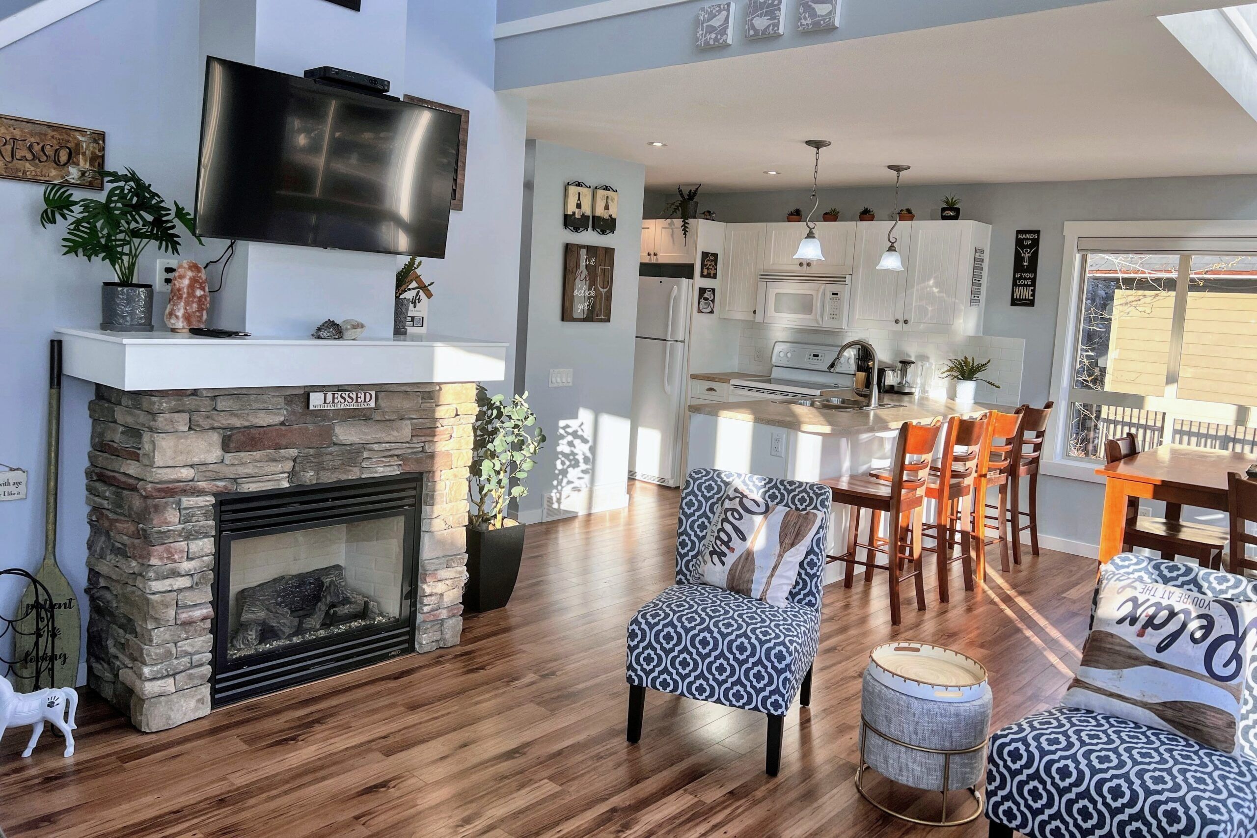 A modern living room with a stone fireplace, a dining area with wooden chairs, and a kitchen with white cabinets and a large TV.
