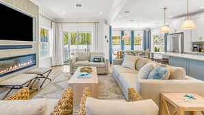 A modern living room with a fireplace, sofas, a coffee table, and a kitchen area with cabinets and a refrigerator. - Popponesset-3-bedrooms, 3.5 baths, sleeps 6, A/C, Pool, Firepit (Mashpee)