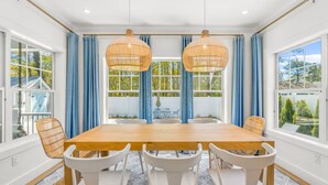 A dining room with a wooden table and white chairs, large windows with blue curtains, and wicker pendant lights. - Popponesset-3-bedrooms, 3.5 baths, sleeps 6, A/C, Pool, Firepit (Mashpee)