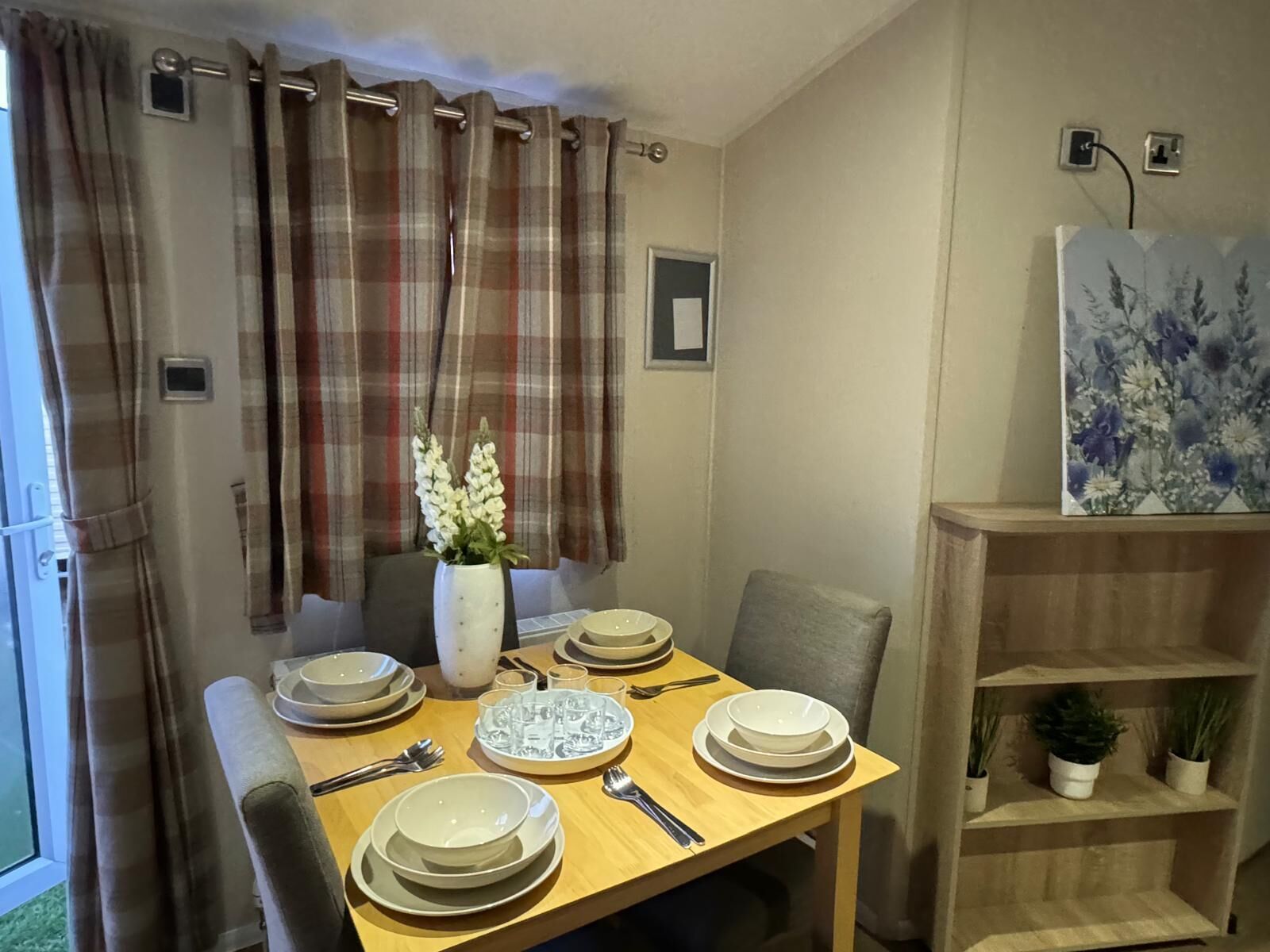A dining area with a wooden table set for four, a vase with white flowers, plaid curtains, and a wall-mounted painting.