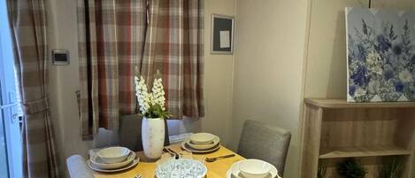 A dining area with a wooden table set for four, a vase with white flowers, plaid curtains, and a wall-mounted painting.
