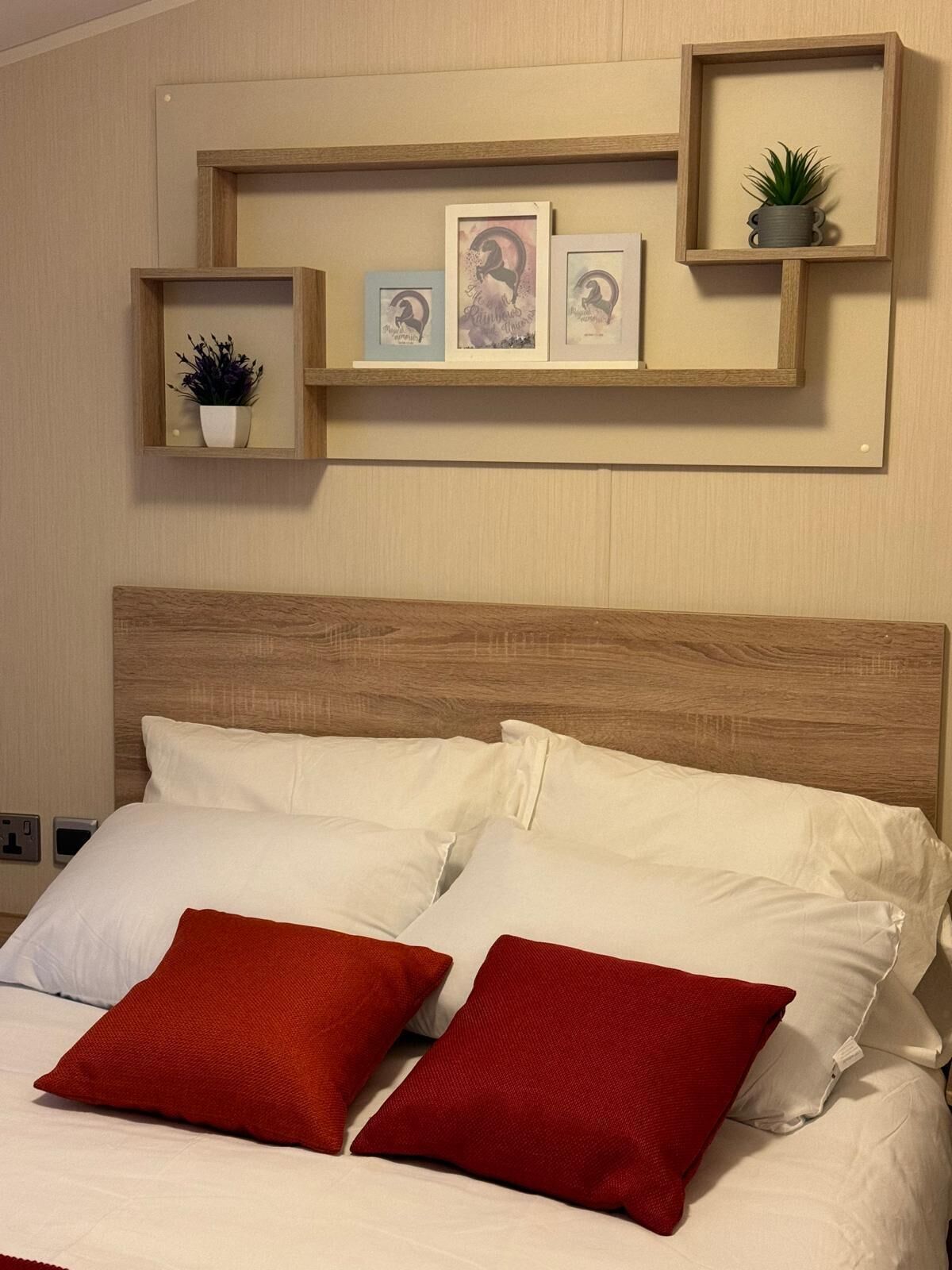A hotel room with a wooden headboard, white bedding, and red pillows. There are wall-mounted shelves with framed pictures and a small potted plant.