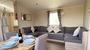 A cozy living area with a sofa, a dining table set with plates and cutlery, and a bottle of wine. - Modern & Blissful Beach Holiday Park Home in Essex (Harwich)