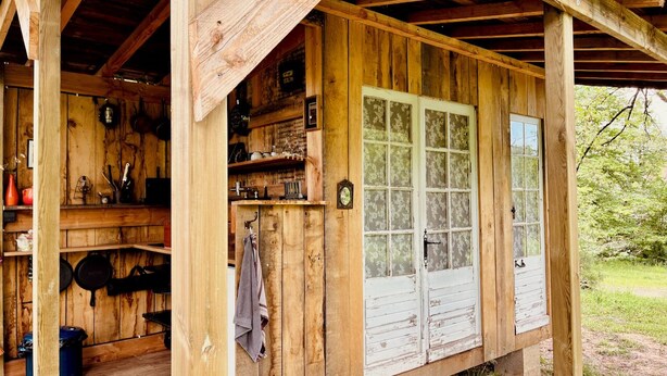 Eine rustikale Holzhütte mit Veranda, einer Küchenzeile mit hängenden Töpfen und einem großen Fenster.
