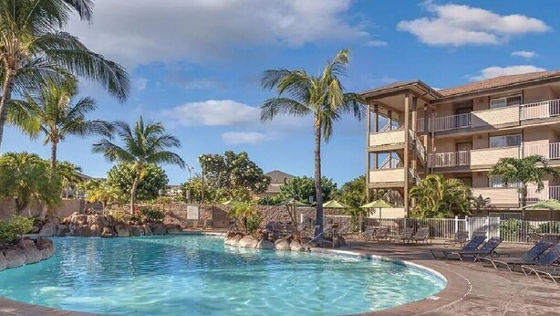 A pool area with lounge chairs, palm trees, and a multi-story building with balconies.