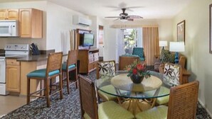 A compact hotel room with a kitchenette, a dining area with a glass table, and a seating area with a ceiling fan.