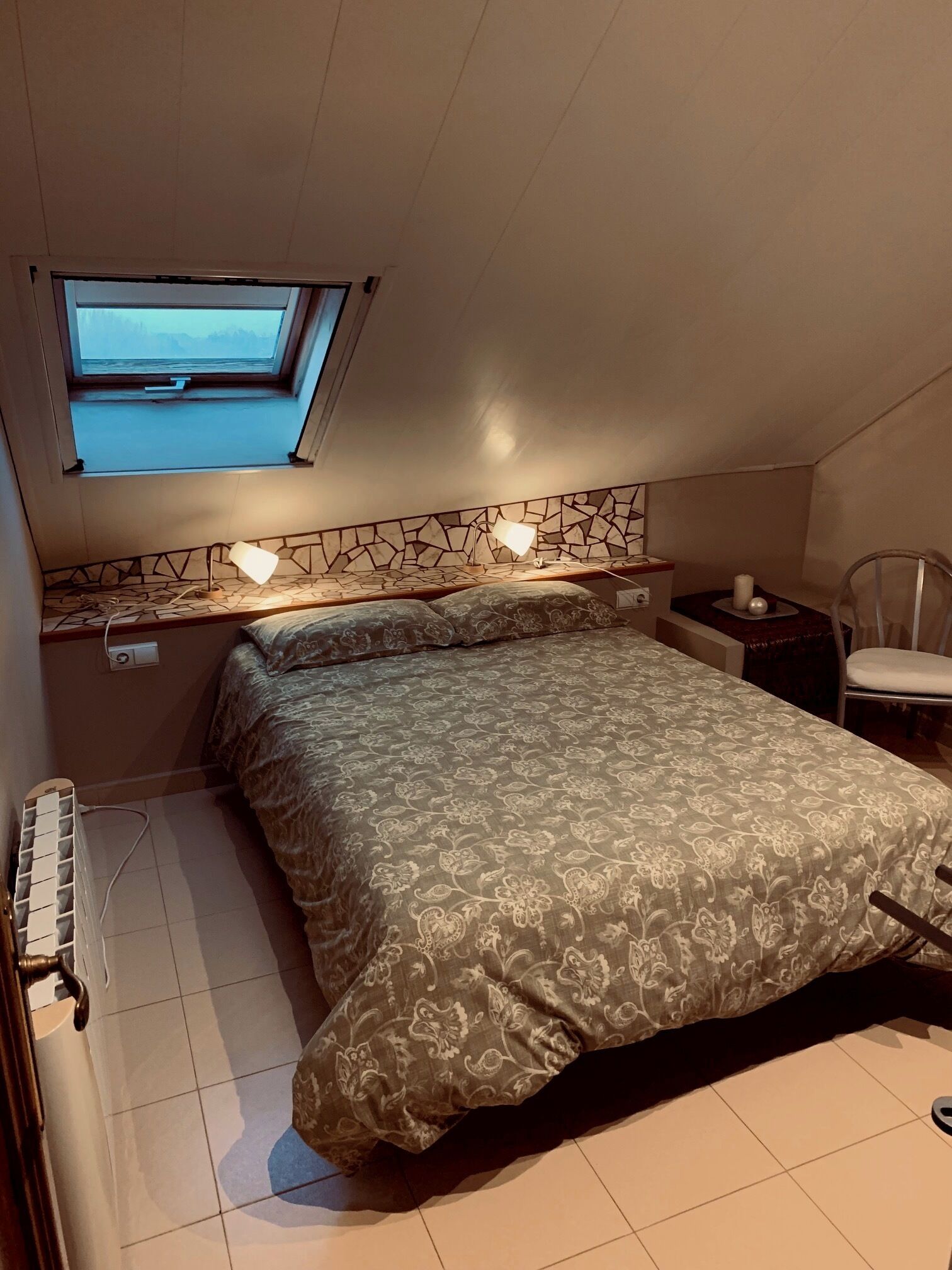 Un dormitorio con cama, una mesita, una silla, un ventanal y un radiador.