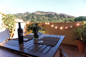 Una mesa de madera con una botella de vino y dos copas en una terraza con vistas a las colinas.