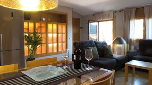 A living room with a sofa, a dining table with a bottle of wine and a glass, and a large window with curtains. - La Terrasseta. Ático con terraza. (Graus)