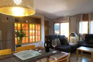 Una sala de estar con un sofá, una mesa de comedor con una botella de vino y una copa, y un ventanal con cortinas.
