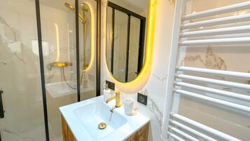 Une salle de bain moderne avec un lavabo blanc, des robinetteries dorées, un grand miroir avec un cadre doré et une cabine de douche en verre.