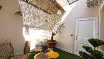 Un petit espace patio avec une table jaune, des chaises en osier, un arbre en pot et une porte blanche.
