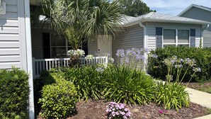 A residential house with a front porch, a small garden with various plants and flowers, and a white picket fence. -  Sunsets on the Sands in the Villages! Golf Front  3/2 (The Villages)