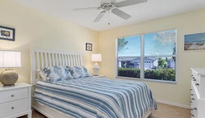 Relax in this serene bedroom with a large bed, nightstand, and dresser, offering a peaceful view from the window.