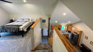 A bedroom with a bed, a nightstand, and a bedside table. There is a staircase leading to a kitchen area with wooden cabinets and a window. - Brightstone Lake Cabin 4 (Rensselaer)