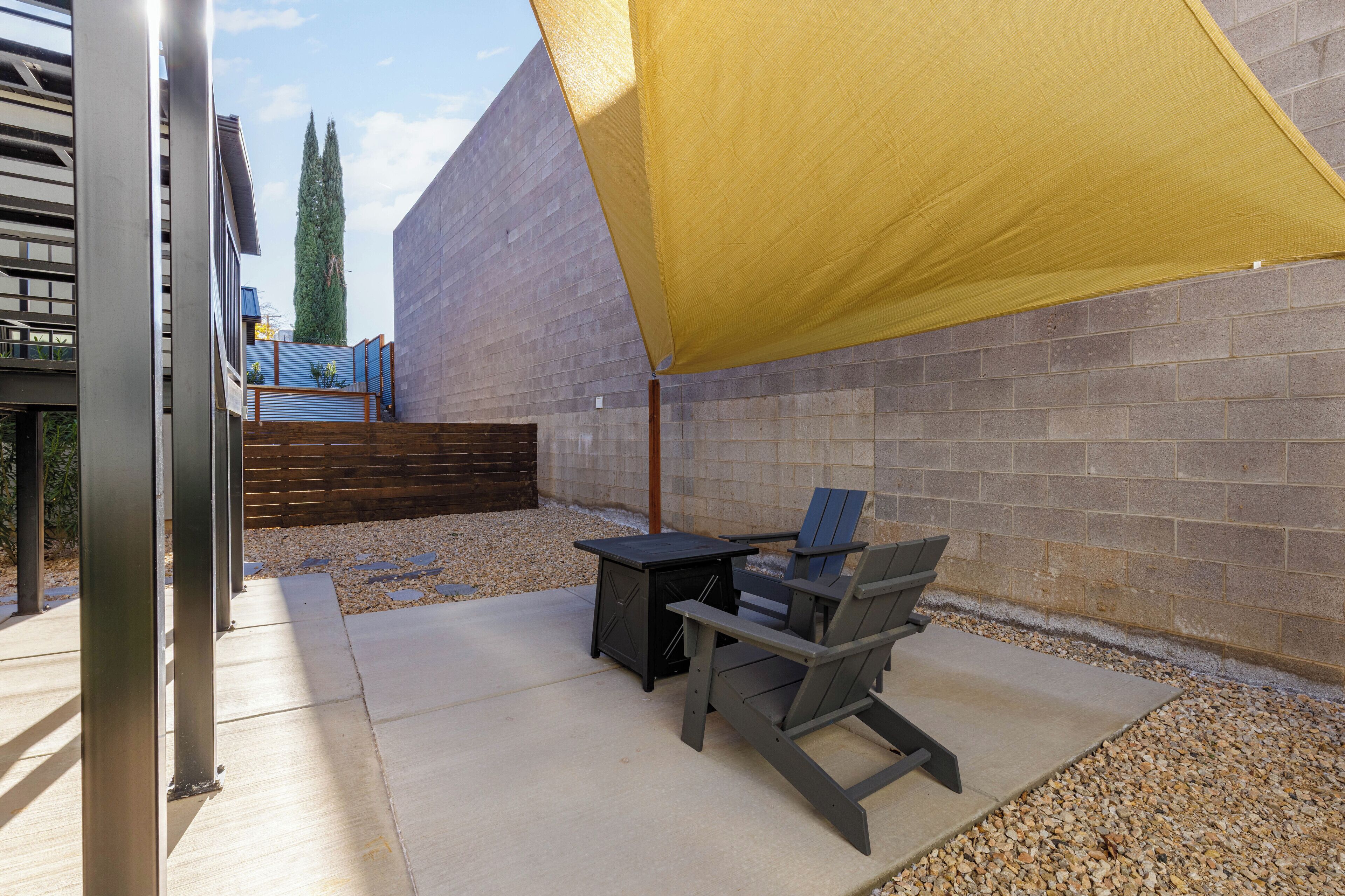 Une terrasse extérieure avec une table et des chaises sous une voile d’ombrage jaune, un mur de briques et une clôture en bois en arrière-plan.