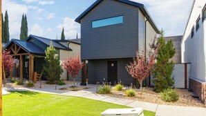 A modern two-story house with a dark exterior, a visible garage door, and a well-maintained front yard with grass and small trees. - Zion Oasis Rentals (Hurricane)