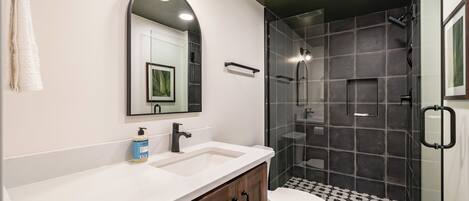 A modern bathroom with a white vanity, a large mirror, a shower with dark tiles, and a toilet.