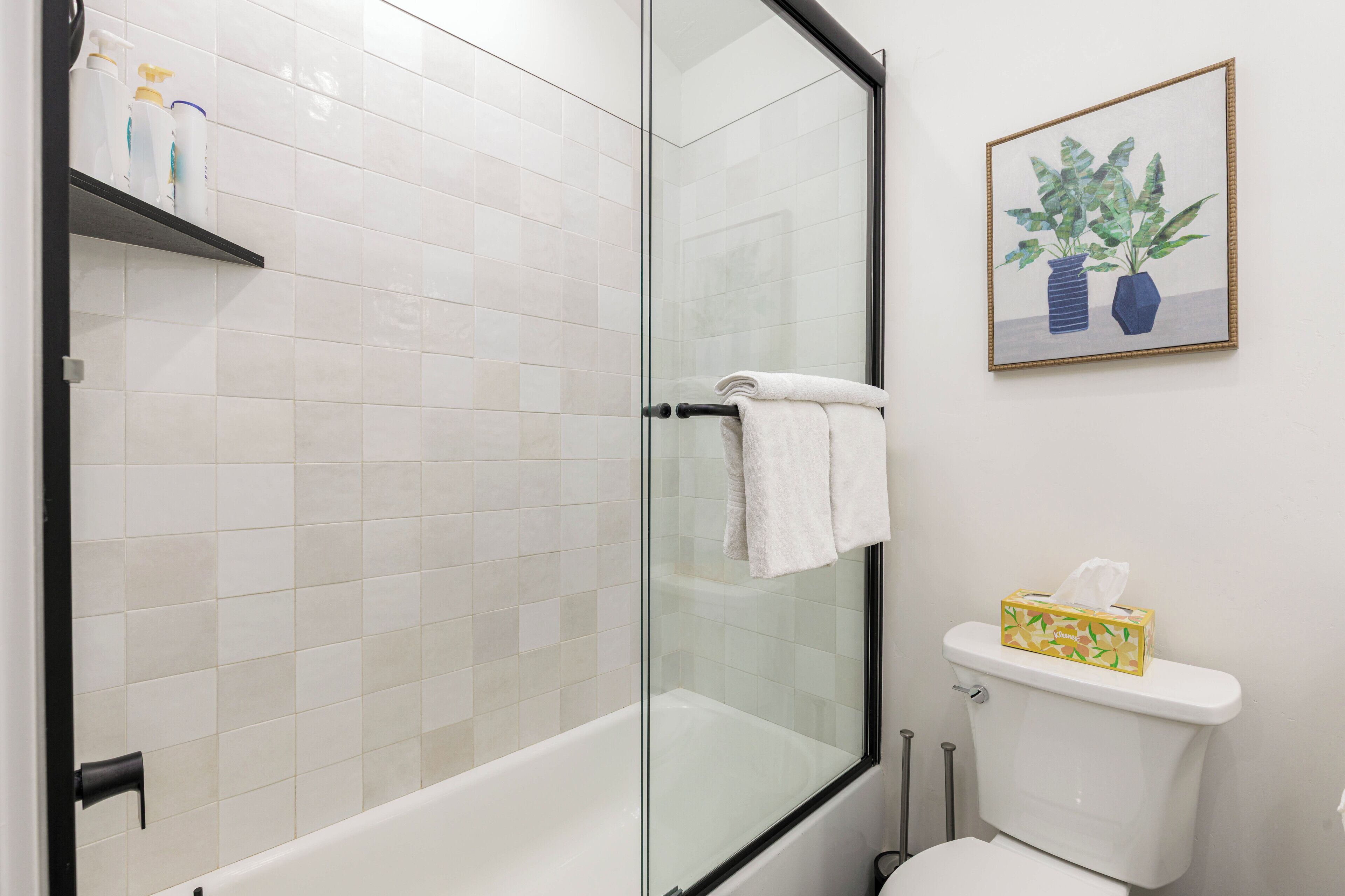 Un baño con ducha envuelta en vidrio, inodoro blanco y repisas con artículos de tocador.
