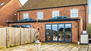 Een bakstenen huis met twee aanbouwen, grote glasramen en een houten omheining.
