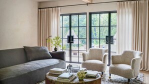 Una sala de estar con un sofá gris, dos sillones, una mesa de centro con libros y un florero, y ventanas grandes con cortinas.