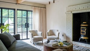 Una sala de estar acogedora con un gran ventanal, dos sillones, un sofá, una chimenea y una mesita de centro con libros y un jarrón.
