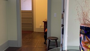 Un pasillo con suelo de madera, una mesa de vidrio con un florero, y una puerta que da a otra habitación.
