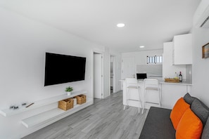 A modern living room with a flat-screen TV, white furniture, and a kitchen area in the background.
