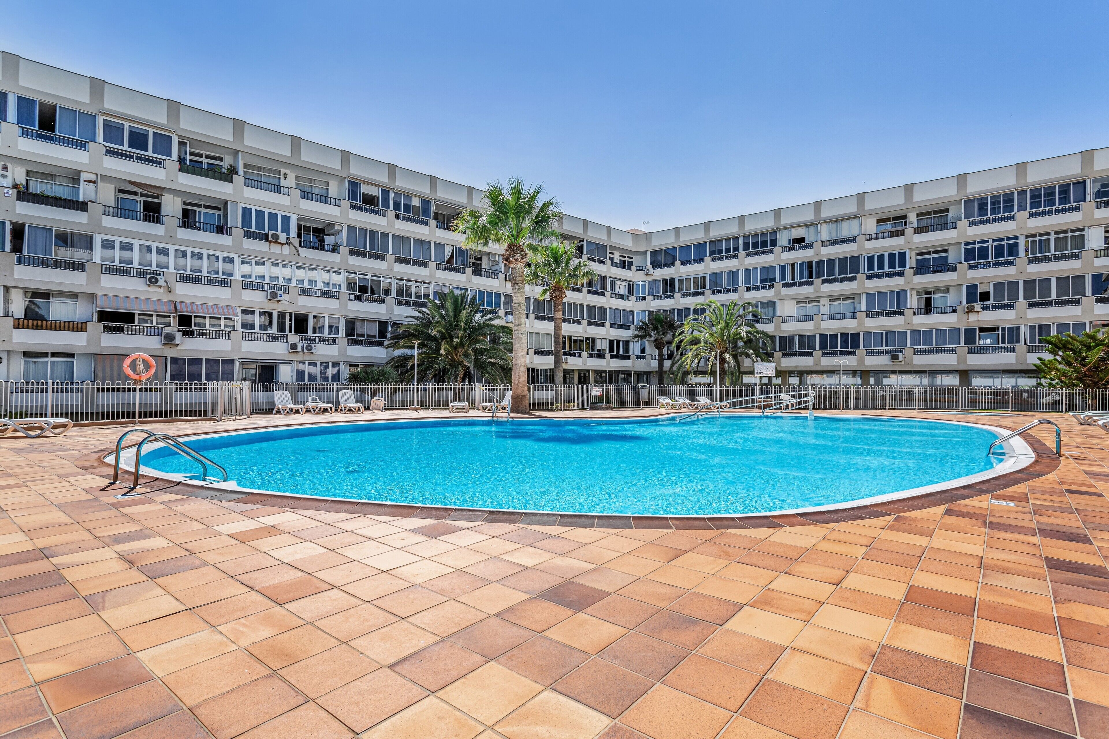 A swimming pool with a tiled deck, surrounded by a multi-story building with balconies and palm trees.