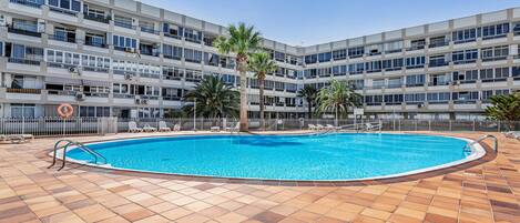 A swimming pool with a tiled deck, surrounded by a multi-story building with balconies and palm trees.