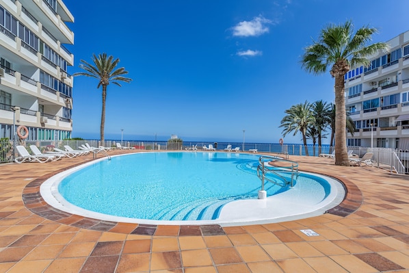 A swimming pool with lounge chairs and palm trees, surrounded by a building with balconies.