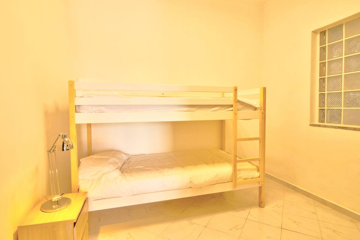 Habitación con literas, una escalera de madera, una mesita de noche con lámpara y una ventana con diseño de cuadrícula.