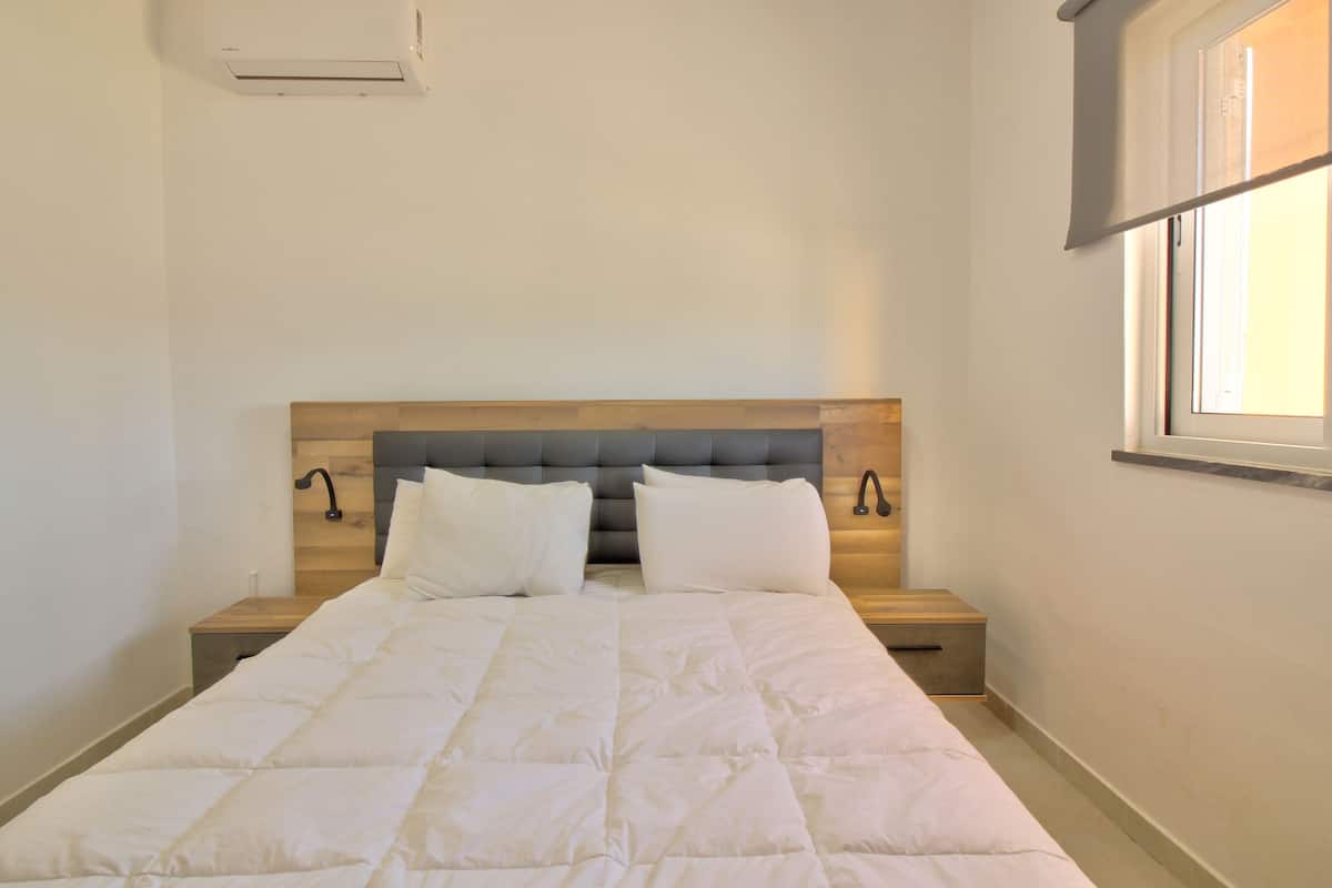 Una cama bien hecha, con cabecera gris y ropa de cama blanca, en una habitación con ventana y aire acondicionado instalado en la pared.