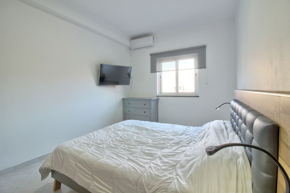 Un dormitorio con cama, cómoda, televisor montado en la pared y ventana.